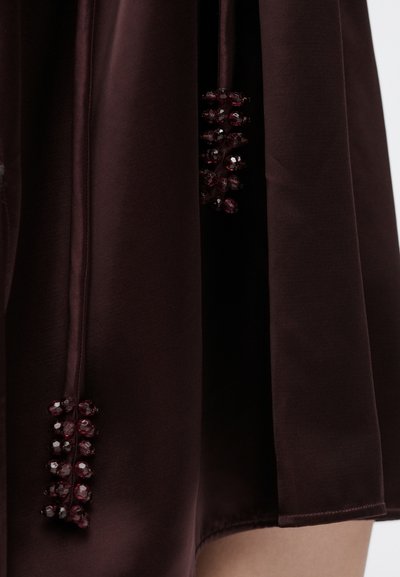 Φόρεμα μπορντό με ομαλή υφή, διακοσμημένο με χάντρες στο τελείωμα. Οι χάντρες σχηματίζουν ένα σύμπλεγμα, προσθέτοντας οπτική λεπτομέρεια.