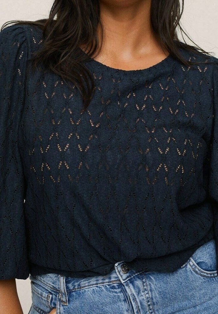 Femme portant un haut bleu marine à motif en dentelle avec des manches bouffantes, associé à un jean taille haute bleu clair.