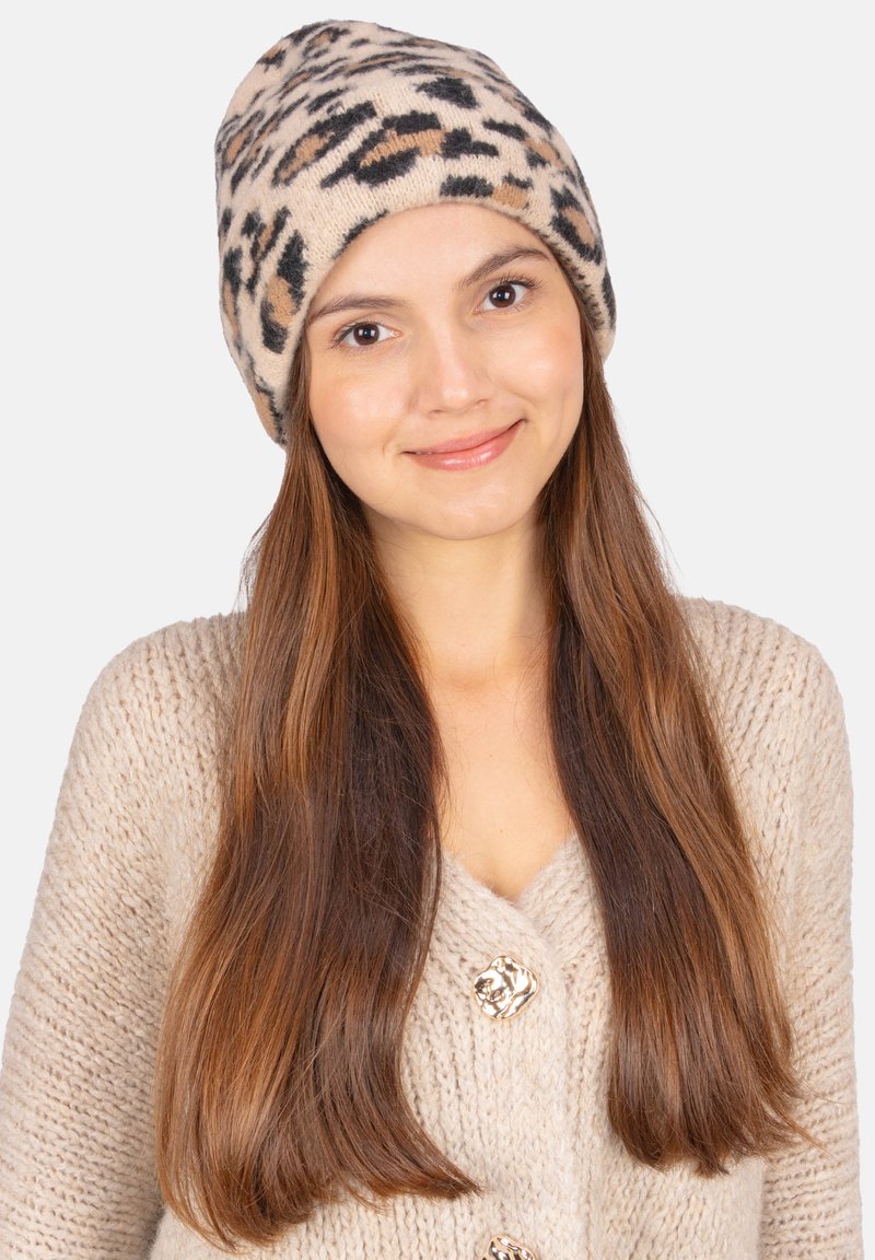 Beanie tricoté beige avec un motif léopard en marron et noir. Le tissu semble doux avec un ajustement serré, complété par un pull de teintes neutres.