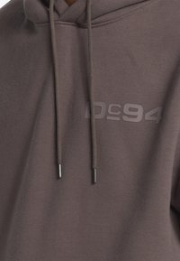 Bruine hoodie van zacht materiaal, voorzien van een logo "DC94" in een lichtere tint aan de voorkant, en tankleurige koorden met metalen punten.