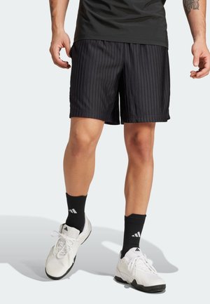 Persoon die zwarte broekjes met krijtstreep draagt, zwarte Adidas-sokken en witte Adidas-sneakers, staand tegen een lichte achtergrond.