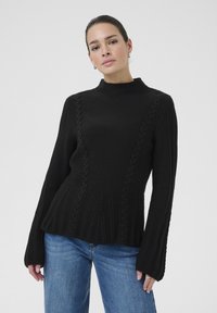 Maglione nero a maglia con colletto alto, motivo testurizzato e maniche a campana. Abbinato a jeans blu, evidenziando una silhouette rilassata.