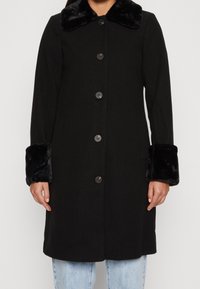Manteau noir avec un col en fausse fourrure et des poignets, présentant un devant à boutons et des manches longues, fabriqué dans un tissu lisse et texturé.