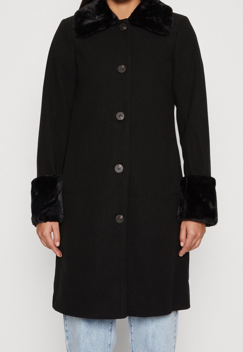 Manteau noir avec un col en fausse fourrure et des poignets, présentant un devant à boutons et des manches longues, fabriqué dans un tissu lisse et texturé.