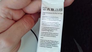 Waschetikett aus weißem Stoff mit Pflegehinweisen in mehreren Sprachen, einschließlich Symbolen für Wasch- und Trockenmethoden, gehalten von einer Hand.