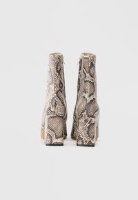 Une paire de bottines avec un motif en peau de serpent beige et noir, des talons carrés et des zips à l'arrière, vue de dos sur un fond blanc.