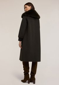 Abrigo de lana verde oscuro con cuello de pelaje sintético, mangas largas y puños acanalados, combinado con botas de piel de ante marrón hasta la rodilla. Textura suave.