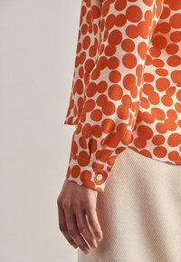 Blusa arancione a pois con maniche lunghe, con polsini imbottiti e realizzata in un tessuto morbido e leggero. Design con colletto e vestibilità comoda.