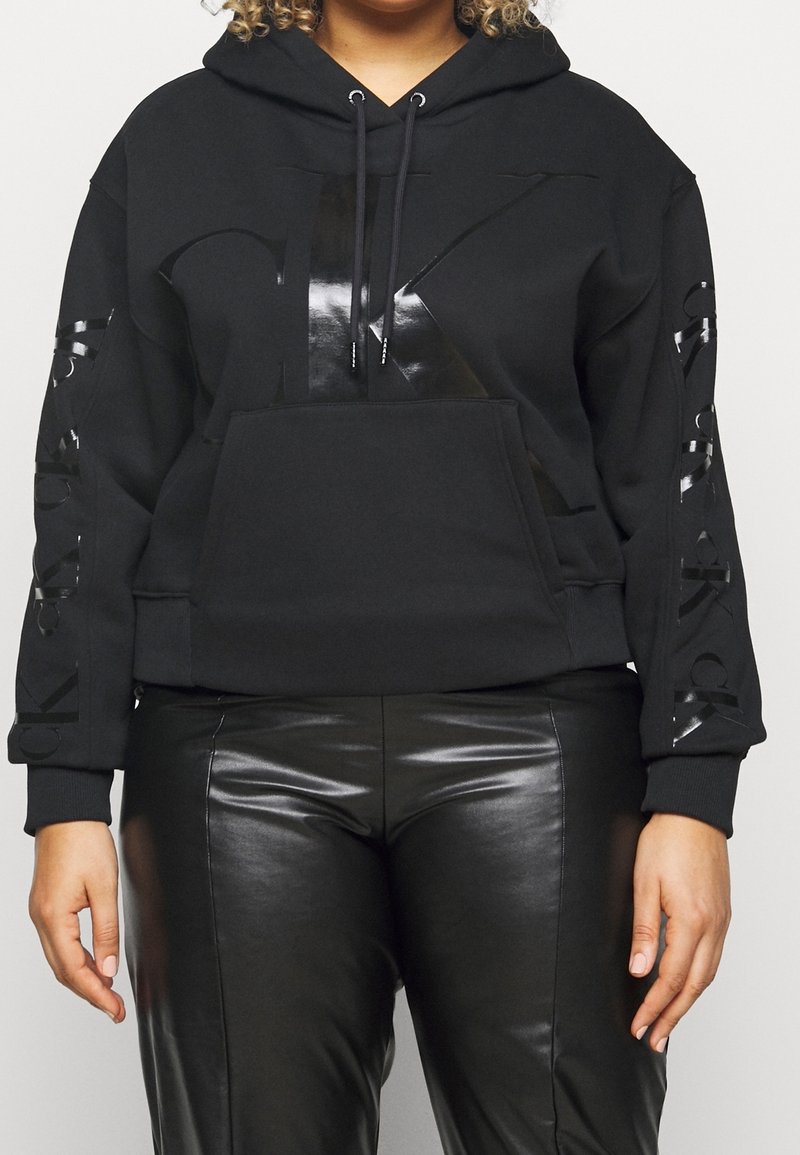 Personne portant un sweat à capuche noir avec des lettres noires brillantes sur la poitrine et les manches, associé à un pantalon en cuir noir.