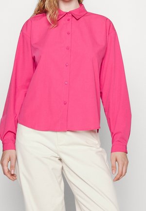 Kobieta ubrana w jaskraworóżową koszulę z długim rękawem na guziki, w połączeniu z jasnobeżowymi spodniami na jednolitym tle.