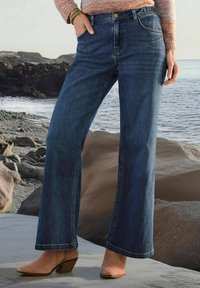 Jeans en denim bleu foncé à jambes larges avec une taille mi-haute, dotés de poches avant et d'une fermeture à bouton. Portés avec des bottines à talons beige.