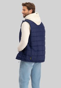 Marineblaues Steppweste mit einem gesteppte Design, darunter ein weißer Kapuzenpullover und hellblaue Jeans. Gelbes Logo-Tag am Saum.