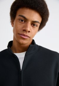 Chaqueta negra con cremallera y cuello alto, hecha de un tejido suave, llevada sobre una camisa blanca. El modelo tiene el cabello corto y rizado y una expresión neutral.