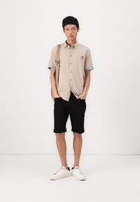 Camisa beige de manga corta con detalles azules y un bolsillo, combinada con pantalones cortos negros y zapatillas blancas. Lleva un bolso bandolera beige.