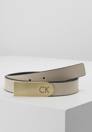 Beżowy skórzany pasek z prostokątną złotą klamrą wygrawerowaną z inicjałami "CK", przedstawiony zwinięty na białej powierzchni.