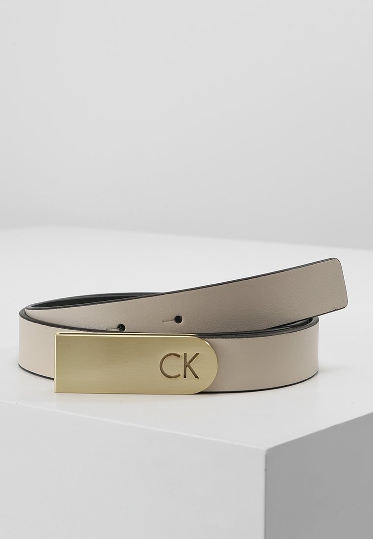 Ceinture en cuir beige avec une boucle rectangulaire en or gravée des initiales "CK", présentée enroulée sur une surface blanche.