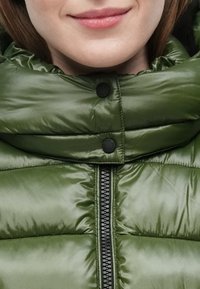 Grøn dunjakke med høj krave, blank overflade, sort lynlås og matchende trykknapper. Nærbillede af stofdetaljer synlige.