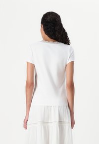 White short-sleeve t-shirt, featuring a lace trim at the neckline, soft cotton material, and a fitted design. Worn with a white, layered skirt.