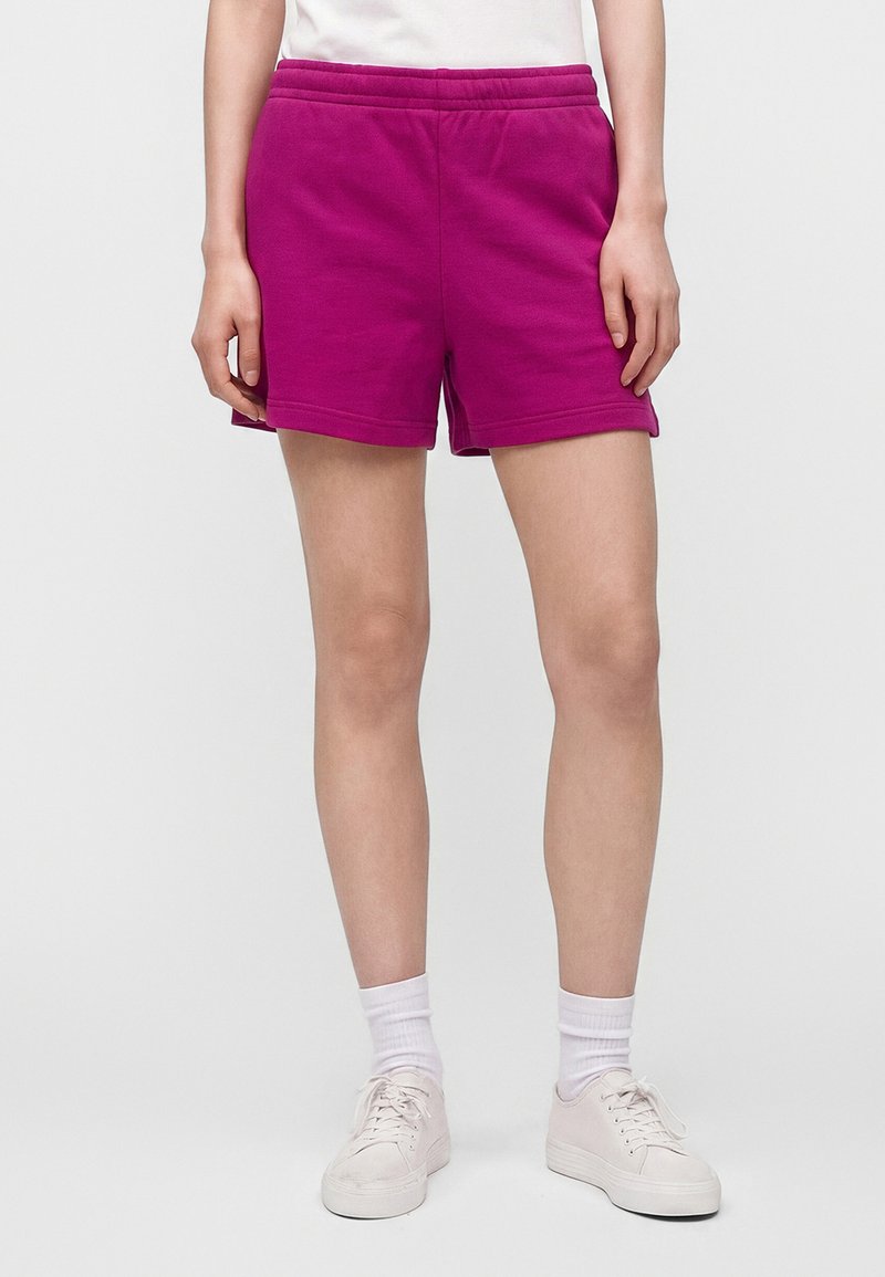 Personne portant un short décontracté rose vif, des chaussettes blanches, des baskets blanches et un haut blanc, debout devant un fond uni.
