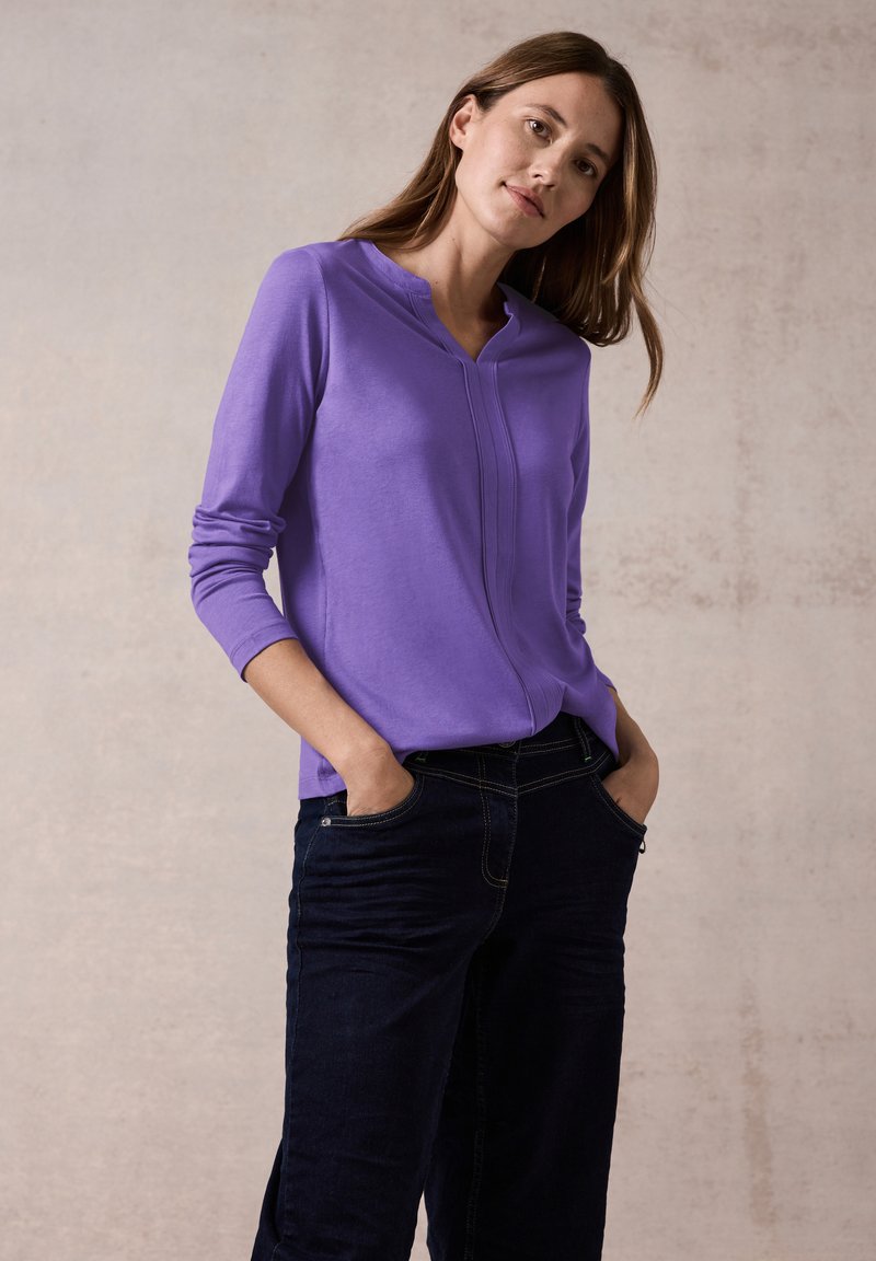 Lila Langarmoberteil mit kragenlosem Design und Nahtdetails an der Vorderseite, kombiniert mit dunkelblauen Jeans, die Taschen und Kontrastnähte aufweisen.
