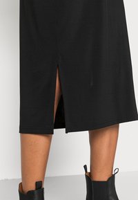 Jupe noire mi-longue avec une fente dans le dos. Fabriquée dans un tissu doux et lisse. Stylisée avec des bottines.