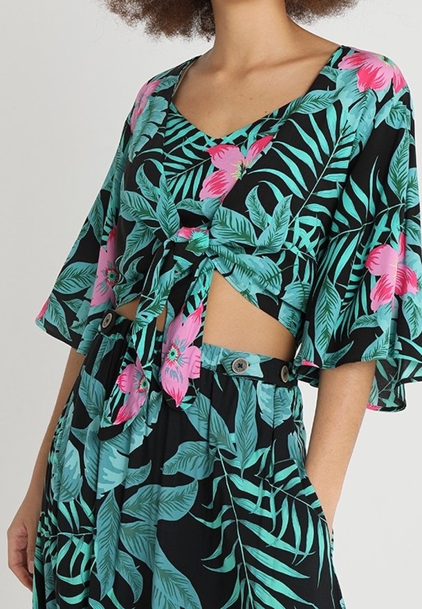 Femme portant une tenue à imprimé tropical avec des feuilles vertes et des fleurs roses, comprenant un crop top noué avec des manches fluides et un pantalon taille haute assorti.