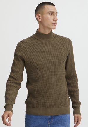 Jersey de tortuga marrón de punto con patrón texturado y puños acanalados. Presenta un corte ajustado y un dobladillo acanalado. Combinado con jeans azules.