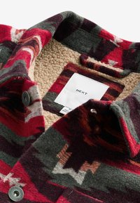 Gros plan sur le col d'une veste en laine rouge et grise à motif, avec une doublure en polaire beige et une étiquette blanche indiquant "NEXT" à l'intérieur.