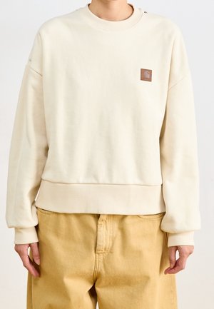 Person iført en cremefarvet sweatshirt med rund hals og en lille brun logo-patch på brystet samt løse beige bukser.