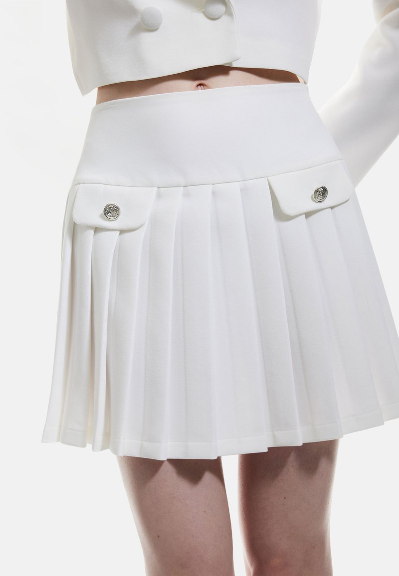 Witte geplooide rok met twee voorzakken, elk voorzien van een zilveren knoop. Soepele stof met een op maat gemaakte tailleband.