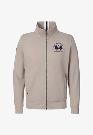 Chaqueta beige con cremallera, cuello alto, dos bolsillos delanteros y logo de jugador de polo azul marino con texto en el pecho izquierdo.