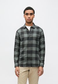 Camisa de flanela aos quadrados verdes e pretos, com mangas compridas, colarinho, botões frontais e bainha curva, usada sobre uma t-shirt branca.