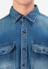 Gros plan d'une personne portant une chemise en denim bleu à boutons avec deux poches poitrine et des coutures visibles.