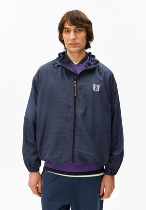 Junger Mann trägt eine marineblaue Zip-up-Jacke mit kariertem Muster und kleinem Aufnäher-Logo, darüber ein lila Sweatshirt.