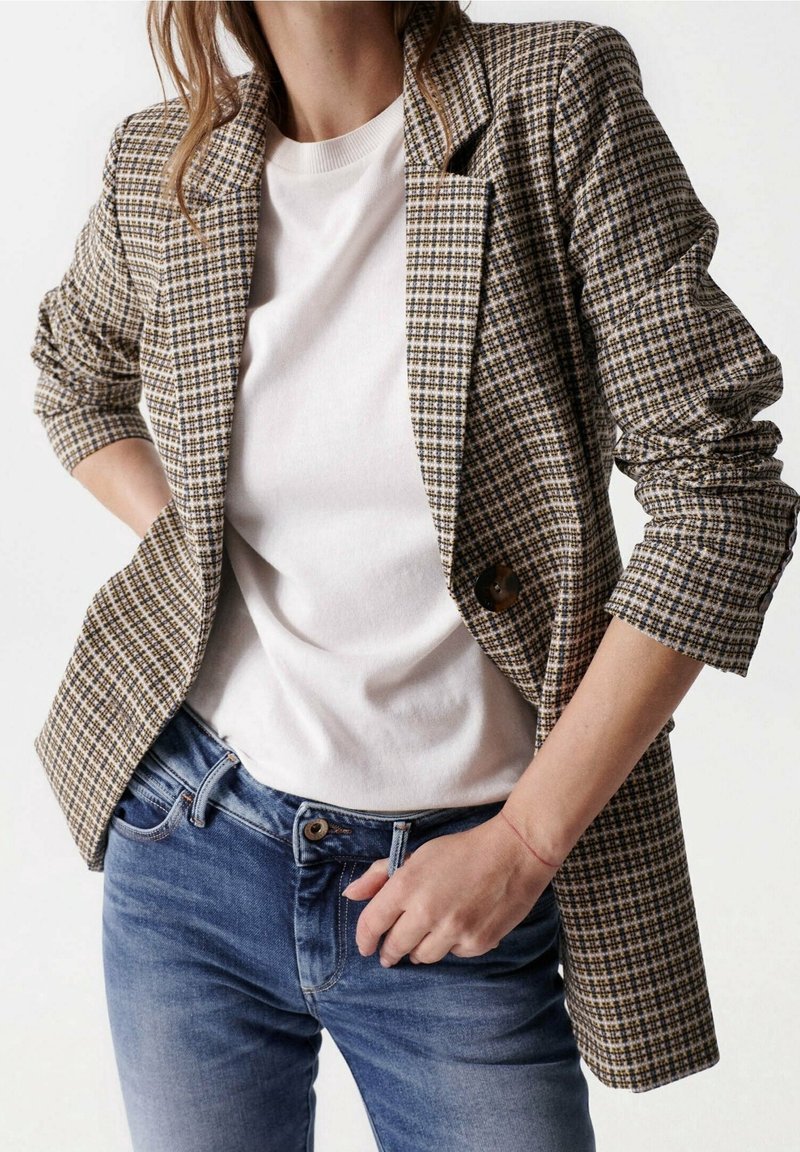 Femme portant un blazer marron à carreaux sur une chemise blanche et un jean bleu, avec une main dans la poche et l'autre ajustant le jean.