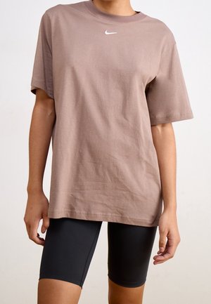 Person trägt ein lockeres braunes Nike-T-Shirt und enge schwarze Radhosen und steht mit entspannten Armen vor einem schlichten Hintergrund.