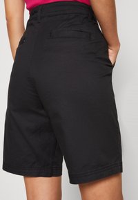 Zwarte korte broek van stof met een gladde textuur. Ze hebben twee achterzakken en een rechte snit die net boven de knie eindigt.