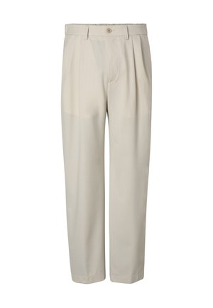 Homme portant un pantalon plissé beige, les mains dans les poches, assorti à des chaussures en daim beige à enfiler et une chemise blanche rentrée.