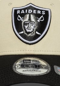 Boné ajustável com parte superior em bege e aba preta, apresentando um logo da Raiders em preto e prata bordado na frente.