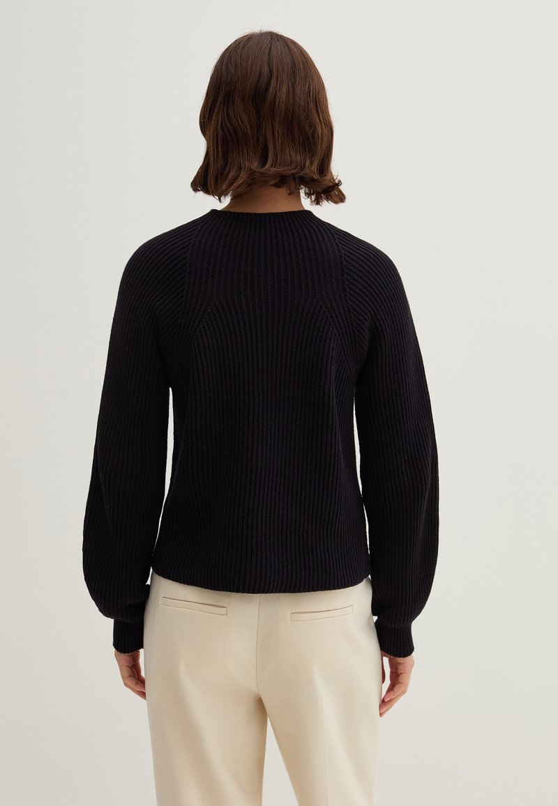 Schwarzer gerippter Strickpullover mit Rundhalsausschnitt und langen Ärmeln, der mit strukturiertem Stoff und einer figurschmeichelnden Silhouette ausgestattet ist, kombiniert mit beigen Hosen.
