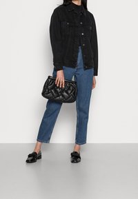 Persona che indossa un giubbotto di denim nero, jeans blu, mocassini neri in pelle lucida, mentre tiene una borsa a tracolla in pelle trapuntata nera su uno sfondo neutro.