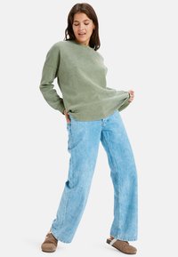 Vrouw in een olijfgroene trui, lichte blauwe wijde jeans en bruine instap schoenen, poseert met één hand in de zak en de andere hand die de zoom van de trui vasthoudt.