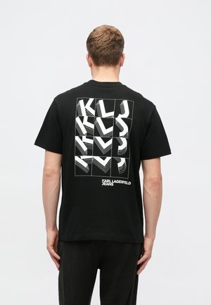 Moški v črni majici s kratkimi rokavi Karl Lagerfeld Jeans, na hrbtni strani z odštevilčenim belim grafičnim besedilom, stoji ob enostavni svetli podlagi.