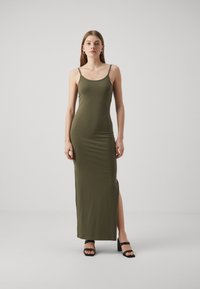 Olivgrön maxiklänning med spaghettiarmstörr, med en sidoöppning, tillverkad av elastiskt tyg. Modellen bär svarta klacksandaler.