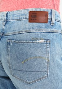 Vaaleansiniset farkut, joissa on nahkainen takapatch, jossa lukee "RAW". Takataskussa on keltaista ommelta ja hieman kulutettu pinta.