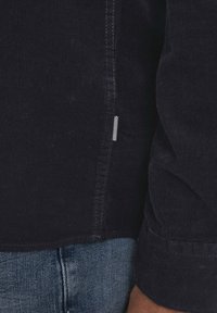 Dark navy corduroy shirt with a ribbed texture, featuring a side hem tag with white and black stripes. Cuffs are neatly finished.