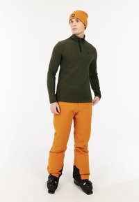 Maglietta verde a maniche lunghe con mezza zip, abbinata a pantaloni arancioni e scarponi da sci neri, indossata da un modello su sfondo bianco.
