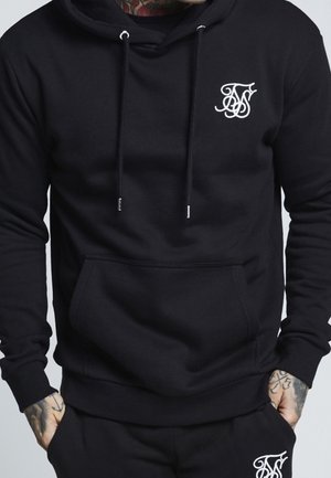Schwarzer Hoodie mit weißem, stilisiertem Logo auf der Brust und passende schwarze Jogginghose, getragen von einer tätowierten Person mit den Händen in den Taschen.