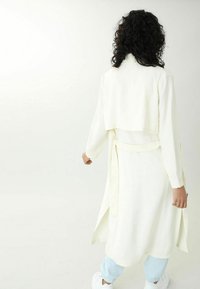 Bílá šaty s dlouhými rukávy, zkráceným přehozem, kimonovým pasem a bočními rozparky, kombinované se světle modrými kalhotami a bílými teniskami.