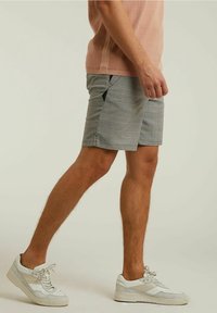 Muž kráčející v kostkovaných šedých kraťasech, světle hnědém pleteném tričku a bílých teniskách na jednobarevném pozadí.